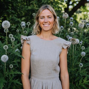 May include: A woman with blonde hair is wearing a light gray linen dress with ruffled sleeves. She is standing in front of a green bush with white flowers.