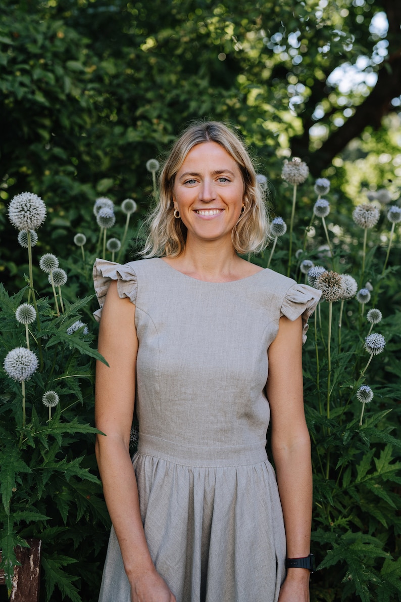 May include: A woman with blonde hair is wearing a light gray linen dress with ruffled sleeves. She is standing in front of a green bush with white flowers.