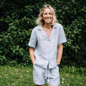 May include: A light blue and white striped linen pajama set. The set includes a short-sleeved button-down shirt and matching shorts with a drawstring waist. The person is smiling and standing in front of a green leafy background.