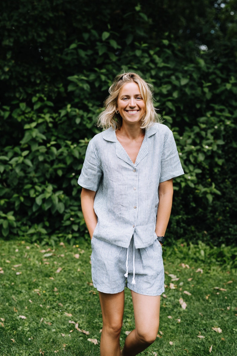 May include: A light blue and white striped linen pajama set. The set includes a short-sleeved button-down shirt and matching shorts with a drawstring waist. The person is smiling and standing in front of a green leafy background.