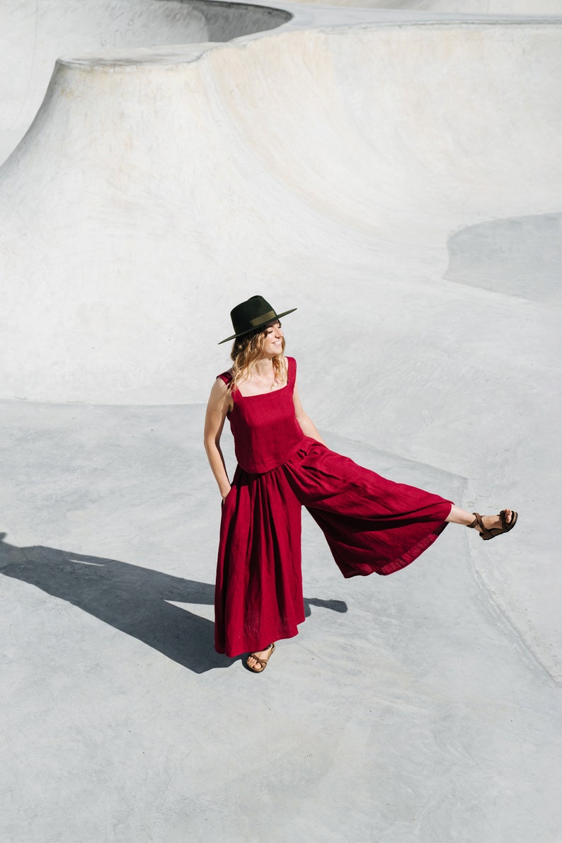 May include: A woman wearing a red linen jumpsuit and a green fedora hat stands in a concrete skate park. She is smiling and has one leg raised.