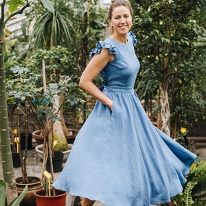 Puede incluir: Una mujer con un vestido de lino azul claro con volantes en las mangas y una falda fruncida se encuentra en un invernadero. Sonr&iacute;e y mira a la c&aacute;mara. El invernadero est&aacute; lleno de plantas y flores.