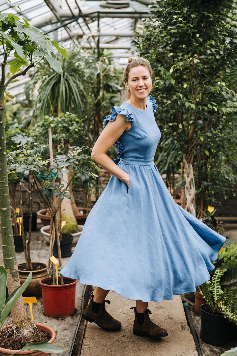 Puede incluir: Una mujer con un vestido de lino azul claro con volantes en las mangas y una falda fruncida se encuentra en un invernadero. Sonr&iacute;e y mira a la c&aacute;mara. El invernadero est&aacute; lleno de plantas y flores.