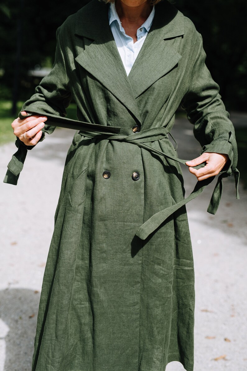 May include: A long, green linen trench coat with a double-breasted closure and a self-tie belt. The coat has a relaxed fit and a slightly flared skirt.