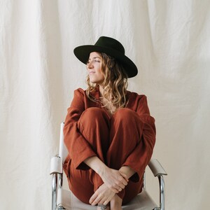 Puede incluir: Una mujer con un mono de color &oacute;xido y un sombrero fedora verde oscuro se sienta en una silla de metal blanco. Tiene el pelo largo y rizado y mira hacia un lado.