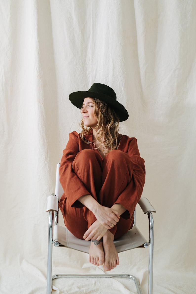 Puede incluir: Una mujer con un mono de color &oacute;xido y un sombrero fedora verde oscuro se sienta en una silla de metal blanco. Tiene el pelo largo y rizado y mira hacia un lado.