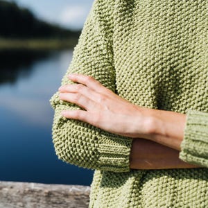 Puede incluir: Primer plano de un suéter de punto verde claro con un patrón texturizado. El suéter tiene cuello redondo y puños y dobladillo acanalados. Las mangas son largas y los brazos están cruzados. El fondo es una vista borrosa de un lago y el cielo.