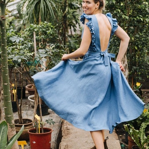 Puede incluir: Una mujer con un vestido de lino azul con la espalda baja y volantes en los hombros est&aacute; de pie en un invernadero. Lleva botas Chelsea marrones y sonr&iacute;e a la c&aacute;mara.