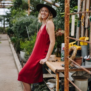 May include: A woman wearing a red dress and a wide-brimmed hat stands in a greenhouse. She is leaning on a wooden table with various gardening tools and plants.