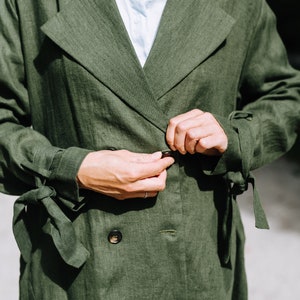 May include: A green linen trench coat with a double breasted closure and tie belt. The coat has a relaxed fit and is made of a lightweight fabric.