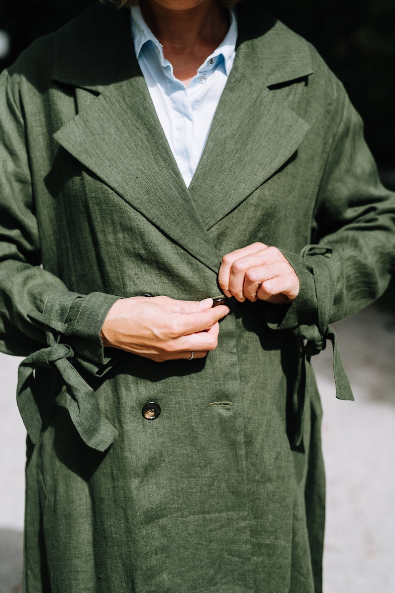 May include: A green linen trench coat with a double breasted closure and tie belt. The coat has a relaxed fit and is made of a lightweight fabric.