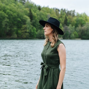 May include: A woman wearing a green linen dress with a black hat stands by a lake. The dress has a button-up front and a tie belt at the waist.