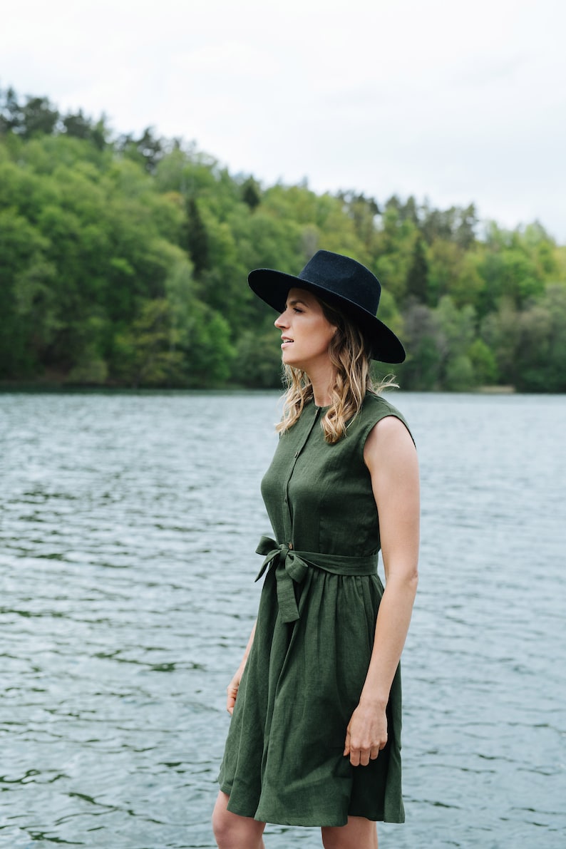 May include: A woman wearing a green linen dress with a black hat stands by a lake. The dress has a button-up front and a tie belt at the waist.