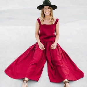 May include: A woman wearing a wide-brimmed green hat, a red linen top and matching wide-leg pants, and brown sandals. She is sitting on a concrete surface.