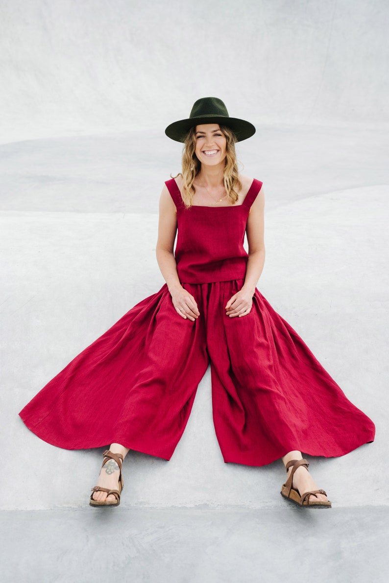 May include: A woman wearing a wide-brimmed green hat, a red linen top and matching wide-leg pants, and brown sandals. She is sitting on a concrete surface.