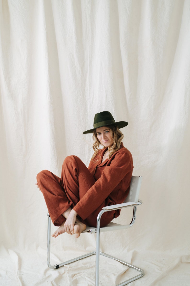 Puede incluir: Una persona con un mono de color &oacute;xido y un sombrero fedora verde oscuro se sienta en una silla de metal blanca sobre un fondo blanco.