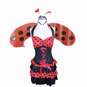 以下が含まれることがあります： 黒いボディスと赤い水玉模様のトップスとスカートが付いたホルターネックドレスが特徴の、てんとう虫のコスチュームセット。コスチュームには、お揃いの羽と触角付きのヘッドバンドが含まれています。ドレスには赤いリボンコルセットが付いています。