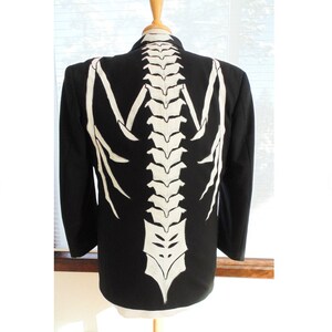 Può includere: Un blazer nero con un disegno di scheletro dipinto di bianco sul retro. Il disegno dello scheletro presenta una colonna vertebrale e costole.