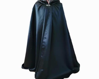 May include: A black hooded cape with a red satin lining. The cape is fastened at the neck with a decorative clasp. The fabric has a slight sheen, suggesting a satin or similar material. The cape is designed to be long and flowing.