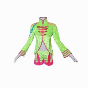 Puede incluir: Un conjunto de disfraz de terciopelo verde brillante con chaqueta y pantalones cortos. La chaqueta tiene charreteras doradas, ribetes rojos y botones decorativos. Los pantalones cortos tienen un diseño rosa en forma de llama. El disfraz se exhibe en un maniquí.