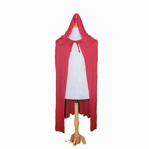 May include: A long, red hooded cape with a tie closure at the neck. The cape is displayed on a white dress form with a wooden stand, set against a white background. The cape's fabric drapes down to the floor.