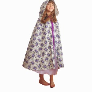 May include: A young girl wearing a white hooded cape with a purple floral pattern and a purple ribbon. The cape has a white fur trim around the hood.