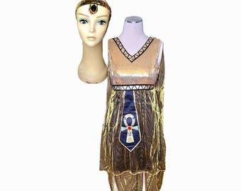 Peut inclure: Robe dorée à paillettes de style égyptien avec un symbole ankh bleu et blanc. Un bandeau doré assorti avec une pierre sombre est également présent. La robe a un col en V et une bordure noire. Le costume est présenté sur une tête et un torse de mannequin.