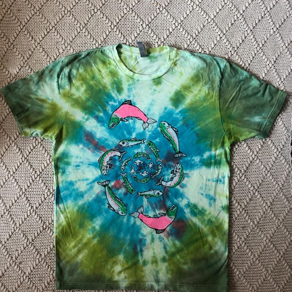 May include: Tie-dye t-shirt with a spiral design featuring fish illustrations. The shirt has a green, blue, and yellow color scheme. The fish are depicted in shades of pink and green.