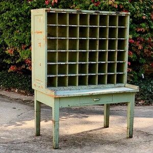 Op de afbeelding: Vintage groene houten postsorteerder met meerdere vakken, een kleine lade en een tafelblad. De verouderde afwerking en de distressed look suggereren een antiek meubelstuk, perfect voor het organiseren van post of kleine items.