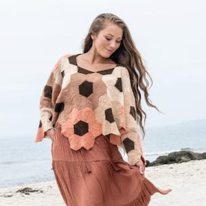 May include: A woman wearing a long, flowing rust-colored skirt and a patterned sweater with floral and geometric designs. The sweater features brown, peach, and cream colors. The outfit is worn on a sandy beach with the ocean in the background.