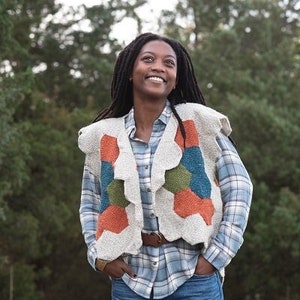 Może przedstawiać: Kobieta w białej dzianinowej kamizelce z kolorowym geometrycznym wzorem, niebiesko-białej koszuli w kratę i niebieskich dżinsach z przetarciami na kolanach. Stoi w lesie.