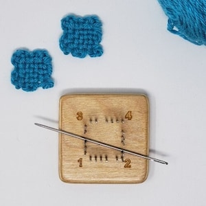 May include: A wooden loom with numbered pegs and a metal needle. Two small blue squares of woven fabric are in the background.