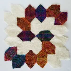 May include: A colourful woven wall hanging with a geometric pattern. The design features a central star shape with eight points, surrounded by smaller shapes in shades of red, orange, blue, green, and purple. The background is white.