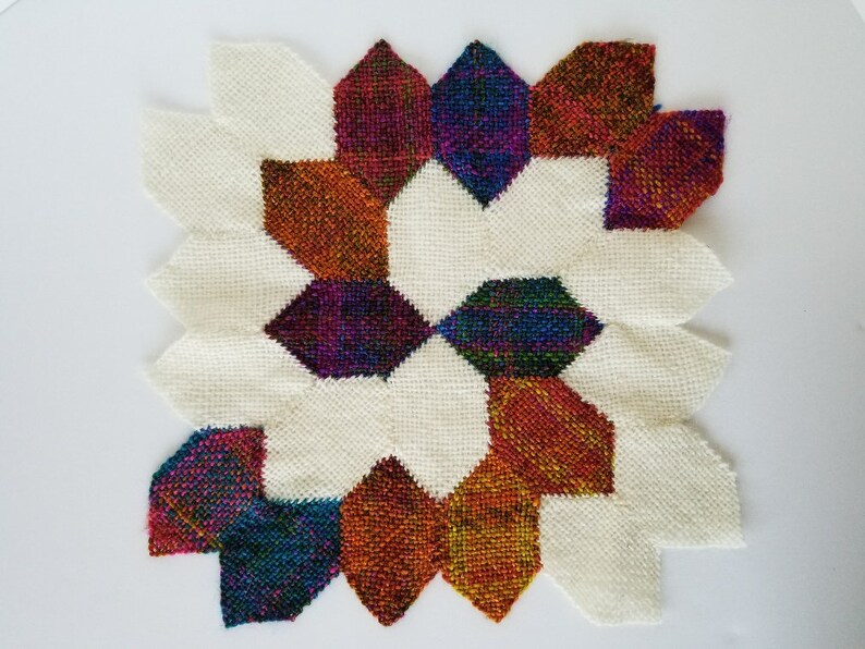 May include: A colourful woven wall hanging with a geometric pattern. The design features a central star shape with eight points, surrounded by smaller shapes in shades of red, orange, blue, green, and purple. The background is white.