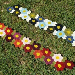 May include: Two crocheted flower-shaped rugs in different colour combinations. The top rug is made with green, blue, and white yarn. The bottom rug is made with red, orange, yellow, and purple yarn.