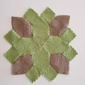 May include: A square knitted coaster with a geometric design. The coaster is made of two colours of yarn: green and brown. The design features eight points, with each point alternating between the two colours.