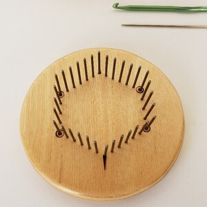 May include: A round wooden loom with metal pegs arranged in a circle. The loom is made of light wood and has a natural finish. The pegs are evenly spaced around the circumference of the loom.