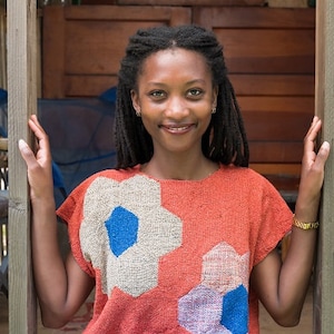 May include: A woman with long black dreadlocks is standing in a doorway. She is wearing a red and white patterned shirt with blue accents. She is smiling and looking at the camera.