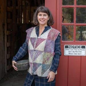 May include: A woman wearing a colorful, patchwork vest with a geometric design. The vest has a zipper and button closure. The woman is standing in front of a red door with a sign that reads, "NOTICE DON'T LET THE ALPACAS OUT, NO MATTER WHAT THEY SAY!"