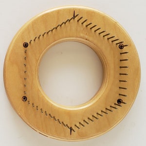 May include: A round wooden knitting loom with metal pegs arranged in a triangular pattern. The loom is light brown and has a natural wood finish.