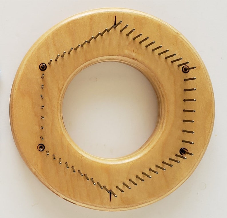 May include: A round wooden knitting loom with metal pegs arranged in a triangular pattern. The loom is light brown and has a natural wood finish.