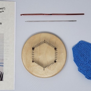 May include: A wooden loom with a hexagonal shape and a blue knitted piece on a white surface. The loom has a row of metal pegs around the edge. The knitted piece is a blue square with a fringe on one side. There are also two knitting needles and a crochet hook on the surface.