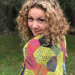 May include: A young person with curly blonde hair wearing a colorful crocheted shawl with a geometric pattern. The shawl is made of different shades of pink, yellow, blue, and green.