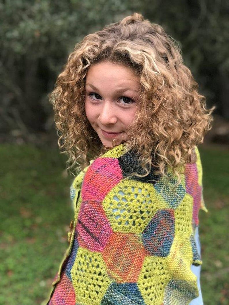 May include: A young person with curly blonde hair wearing a colorful crocheted shawl with a geometric pattern. The shawl is made of different shades of pink, yellow, blue, and green.