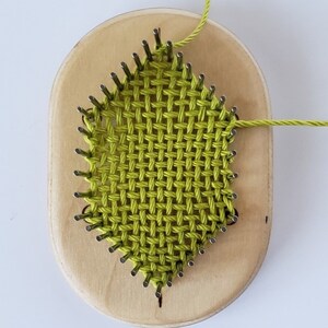 May include: A green yarn woven on a wooden loom with metal pegs. The loom is oval shaped and has a light wood finish.