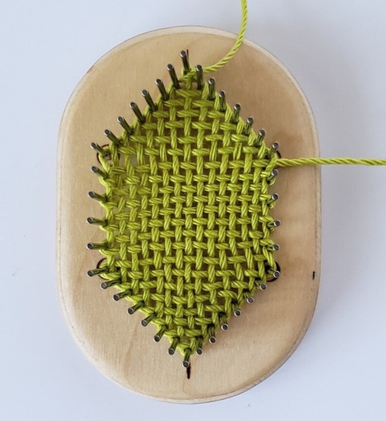 May include: A green yarn woven on a wooden loom with metal pegs. The loom is oval shaped and has a light wood finish.