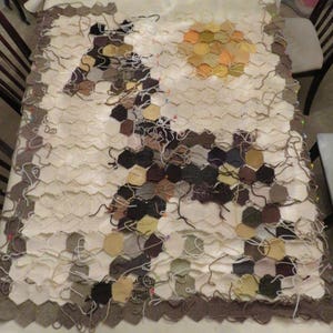 May include: A large, unfinished knitted blanket with a honeycomb pattern. The blanket is made of various shades of brown, beige, and white yarn. The blanket is pinned together with safety pins.