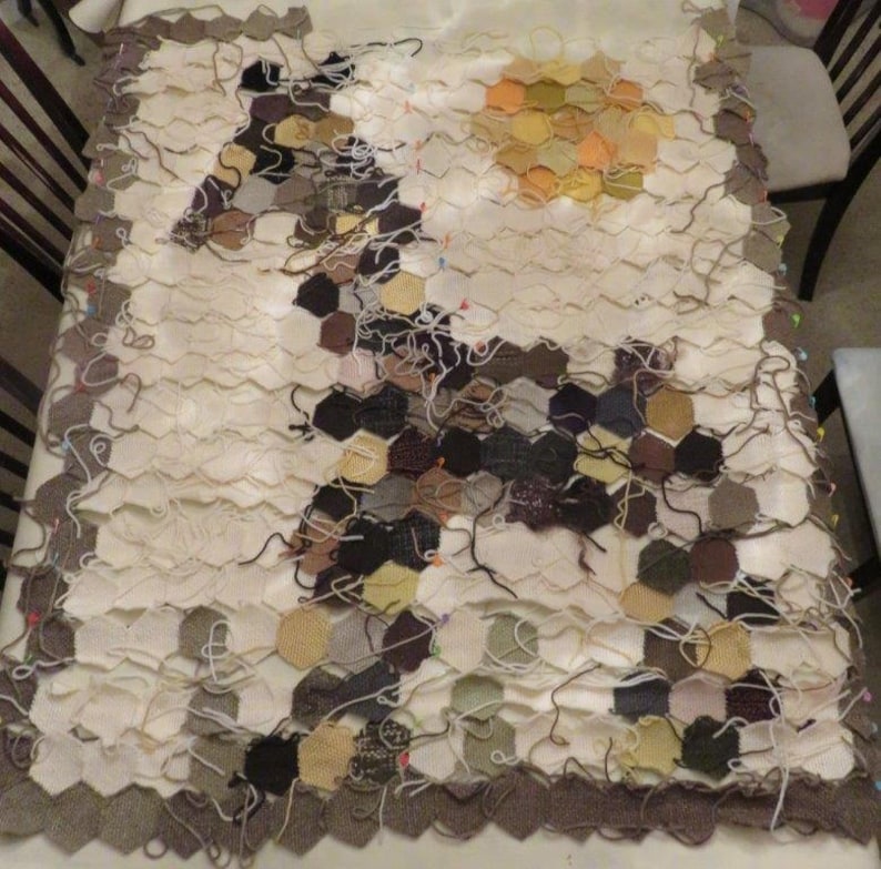 May include: A large, unfinished knitted blanket with a honeycomb pattern. The blanket is made of various shades of brown, beige, and white yarn. The blanket is pinned together with safety pins.