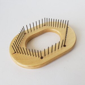 May include: A wooden knitting loom with a rounded oval shape and metal needles. The loom is light brown and has a hole in the centre.