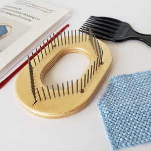 May include: A wooden knitting loom with a rounded shape and a central opening. The loom has rows of metal pegs and is accompanied by a red crochet hook, a black comb, and two knitted squares in blue and green.